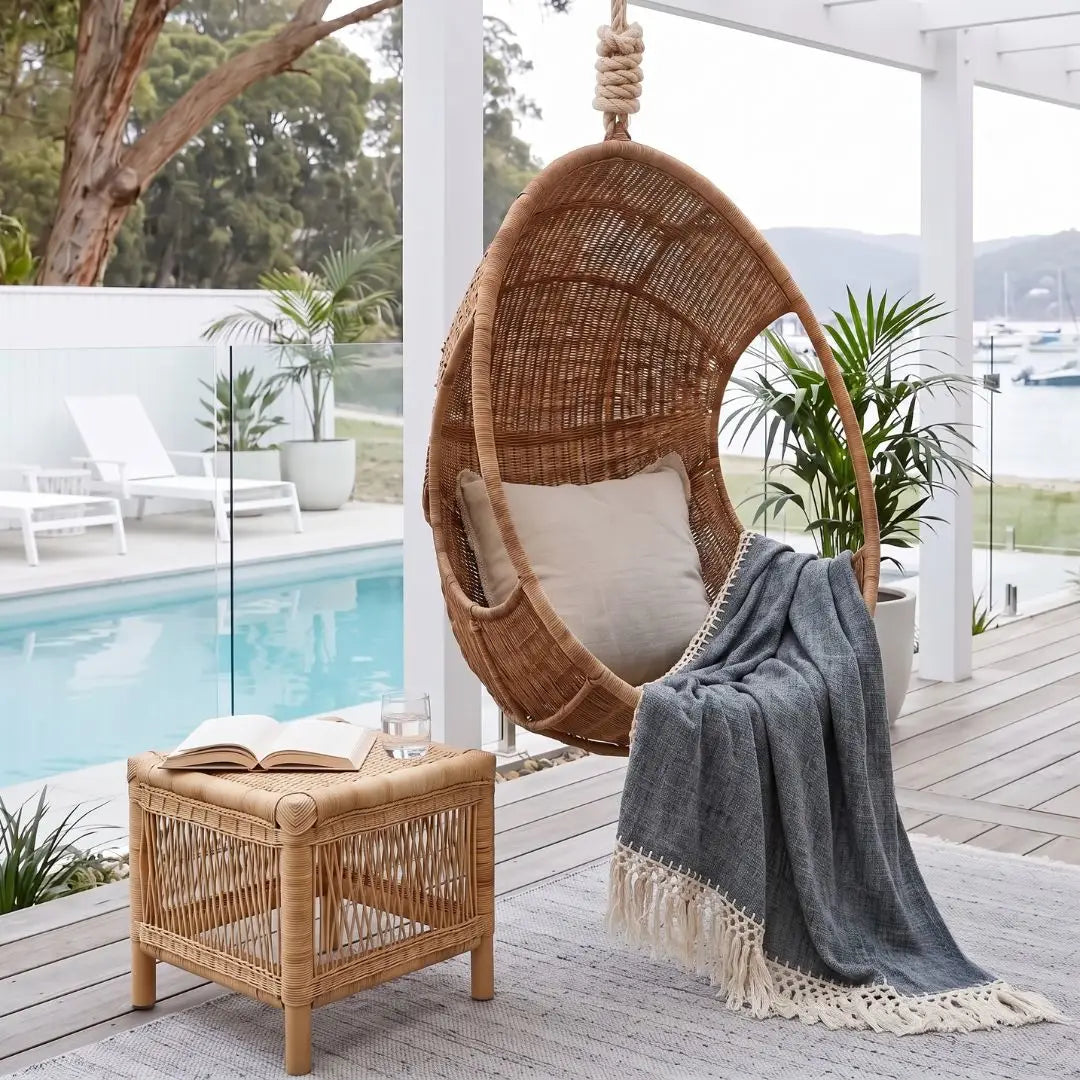 A woven hanging egg chair with a cream cushion and a gray throw blanket, set on a wooden deck beside a pool. A matching woven side table with an open book is in the foreground, with plants, white lounge chairs, and a waterfront view in the background.