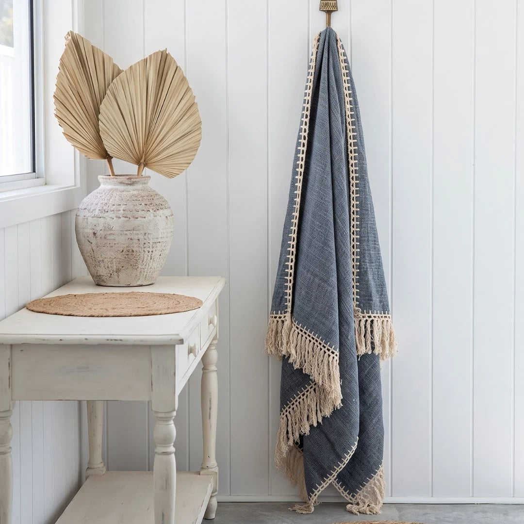 A blue-gray woven throw blanket with cream fringe and border trim hangs from a brass hook on a white paneled wall. To the left, a white side table holds a textured vase with dried palm leaves and a round woven placemat.