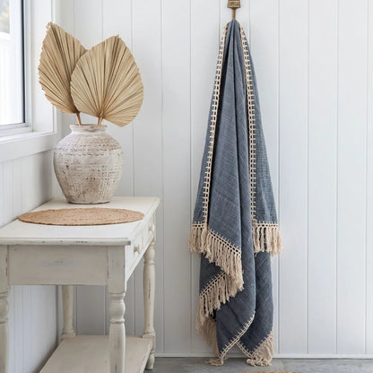 A blue-gray woven throw blanket with cream fringe and border trim hangs from a brass hook on a white paneled wall. To the left, a white side table holds a textured vase with dried palm leaves and a round woven placemat.