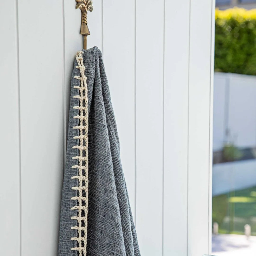 A dark gray textured throw blanket with a cream-colored braided border hangs from a brass hook on a white vertical-panel door, with a blurred outdoor garden background visible on the right.