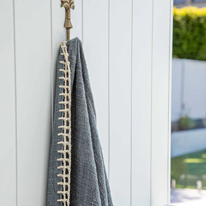 A dark gray textured throw blanket with a cream-colored braided border hangs from a brass hook on a white vertical-panel door, with a blurred outdoor garden background visible on the right.
