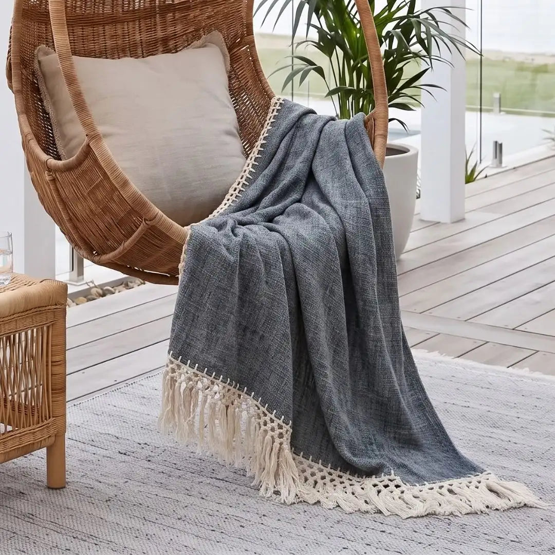 A gray woven throw blanket with cream fringe and crocheted trim draped over a wicker hanging chair on a porch, next to a beige cushion and a potted plant.