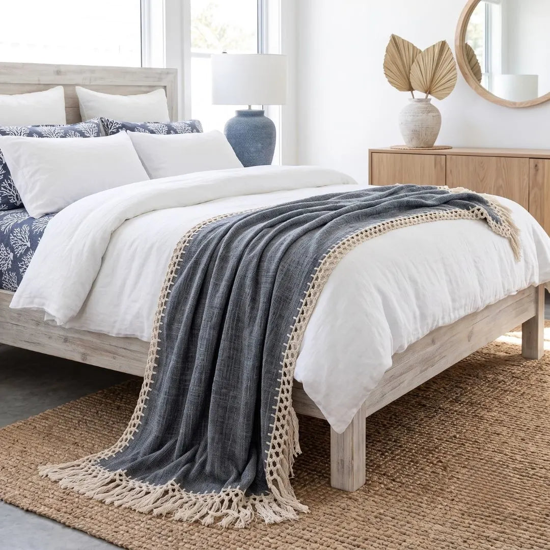 A dark gray textured throw blanket with cream fringe and a braided border draped over a white bed in a bright, modern bedroom.