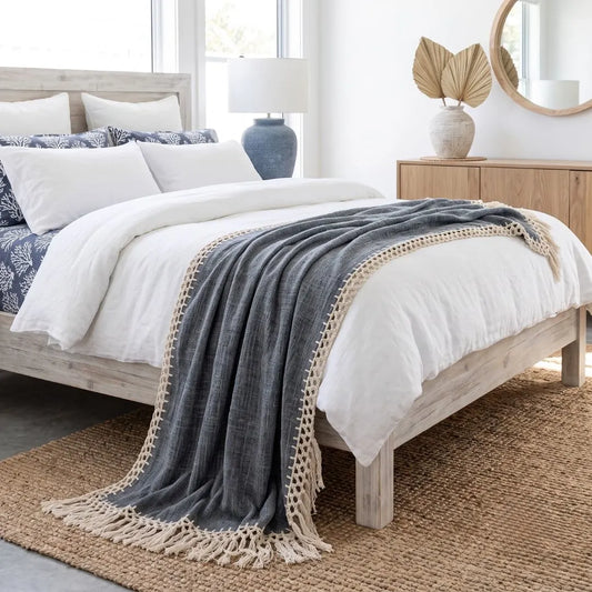 A dark gray textured throw blanket with cream fringe and a braided border draped over a white bed in a bright, modern bedroom.