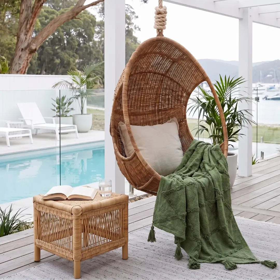 A woven rattan hanging egg chair with a white cushion sits on a covered patio beside a pool, with a green textured throw blanket draped over the seat. A matching woven side table holds an open book and a glass, with plants and a pool visible in the background.