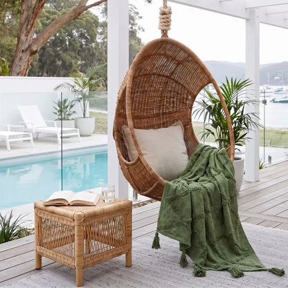 A woven rattan hanging egg chair with a white cushion sits on a covered patio beside a pool, with a green textured throw blanket draped over the seat. A matching woven side table holds an open book and a glass, with plants and a pool visible in the background.