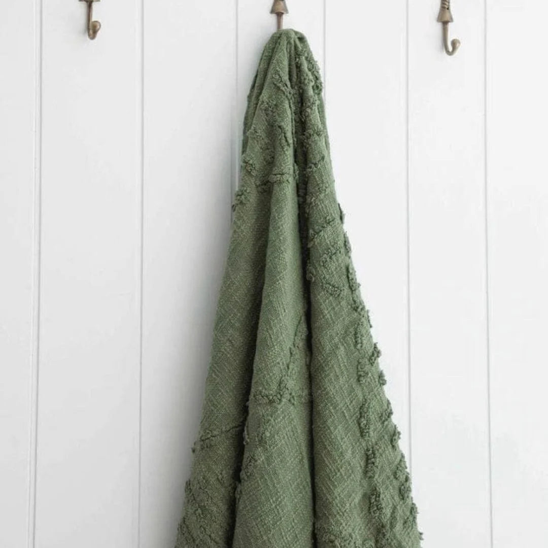 An olive green textured throw blanket with raised tufted patterns hangs from a brass wall hook on a white shiplap wall.
