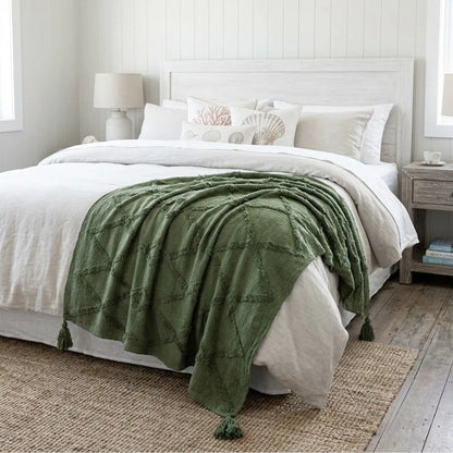 An olive green textured throw blanket with tassel ends draped over a bed with white linens in a bright, modern bedroom setting.