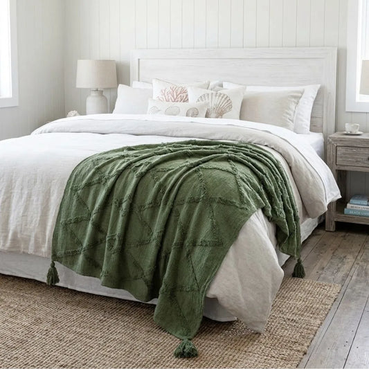 An olive green textured throw blanket with tassel ends draped over a bed with white linens in a bright, modern bedroom setting.