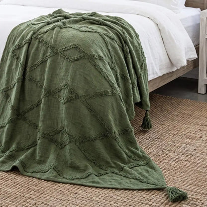An olive green textured throw blanket with a raised geometric diamond pattern and tassels, draped over a bed with white linens on a woven jute rug.