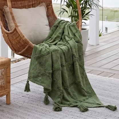 An olive green textured throw blanket with tassel ends draped over a wicker hanging chair on a wooden deck patio.