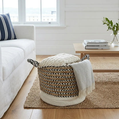 A woven storage basket with black-and-cream striped handles sits on a natural jute rug in a bright living room. A chunky cream cable-knit throw is folded inside the basket, and a light gray fringed throw blanket is draped over the side.