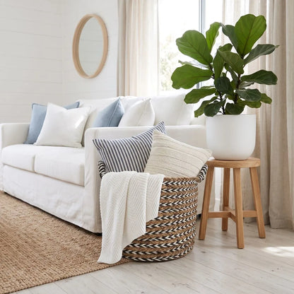A styled living room scene featuring a white sofa with blue and white pillows, a woven basket holding a cream throw blanket, and a large green fiddle-leaf fig plant in a white pot on a small wooden stool.