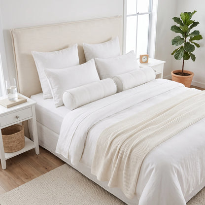 A neatly made bed in a bright bedroom setting, dressed in white bedding with multiple white pillows, a cylindrical bolster pillow, and a cream ribbed throw blanket. The bed has a tall upholstered headboard and is flanked by white bedside tables with small decor items, a window, and a potted green plant.