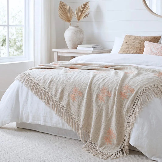 A beige woven throw blanket with coral-pink floral embroidery and fringe trim draped over a white bed in a bright, modern bedroom.