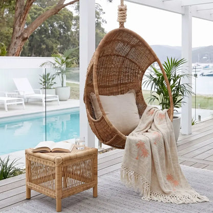 A woven hanging egg chair with a cream cushion sits on a wooden deck beside a small rattan side table with an open book and a cream throw blanket featuring coral palm tree prints. A swimming pool, white lounge chairs, and potted plants are visible in the background.