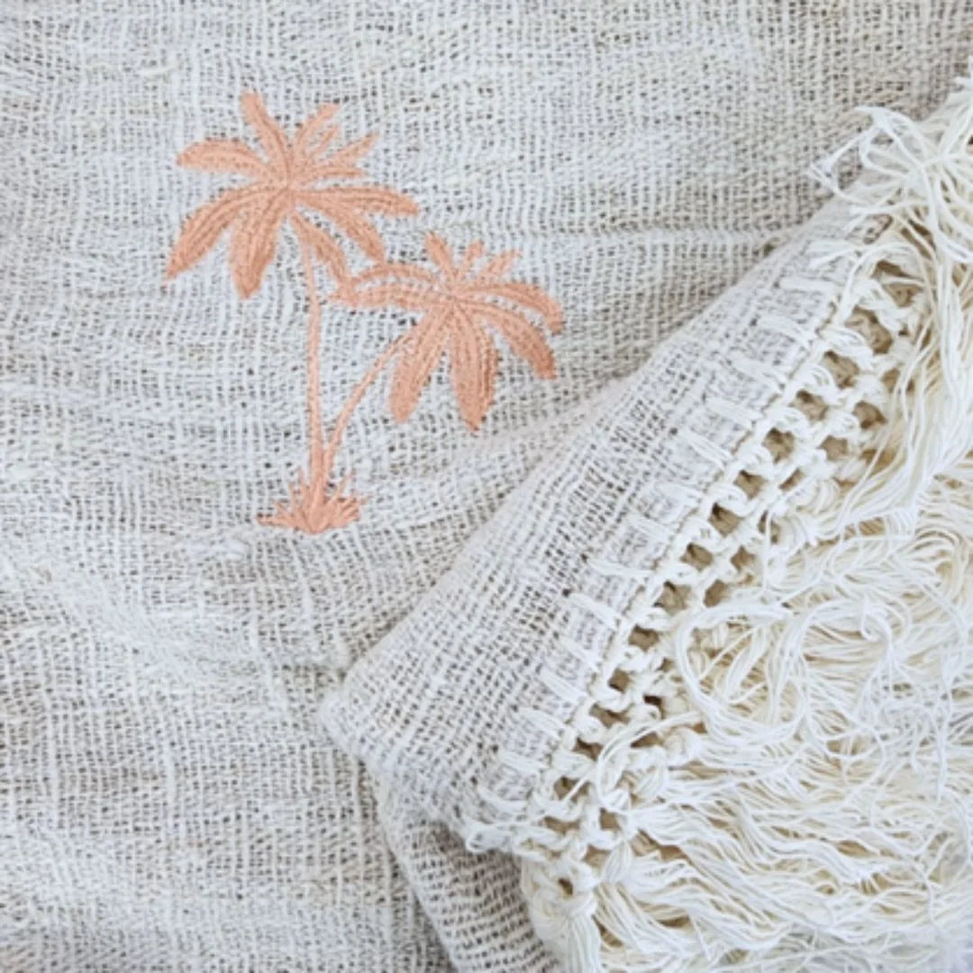 Close-up of a woven textile in warm off-white with peach embroidered palm trees and a cream fringed crochet edge.