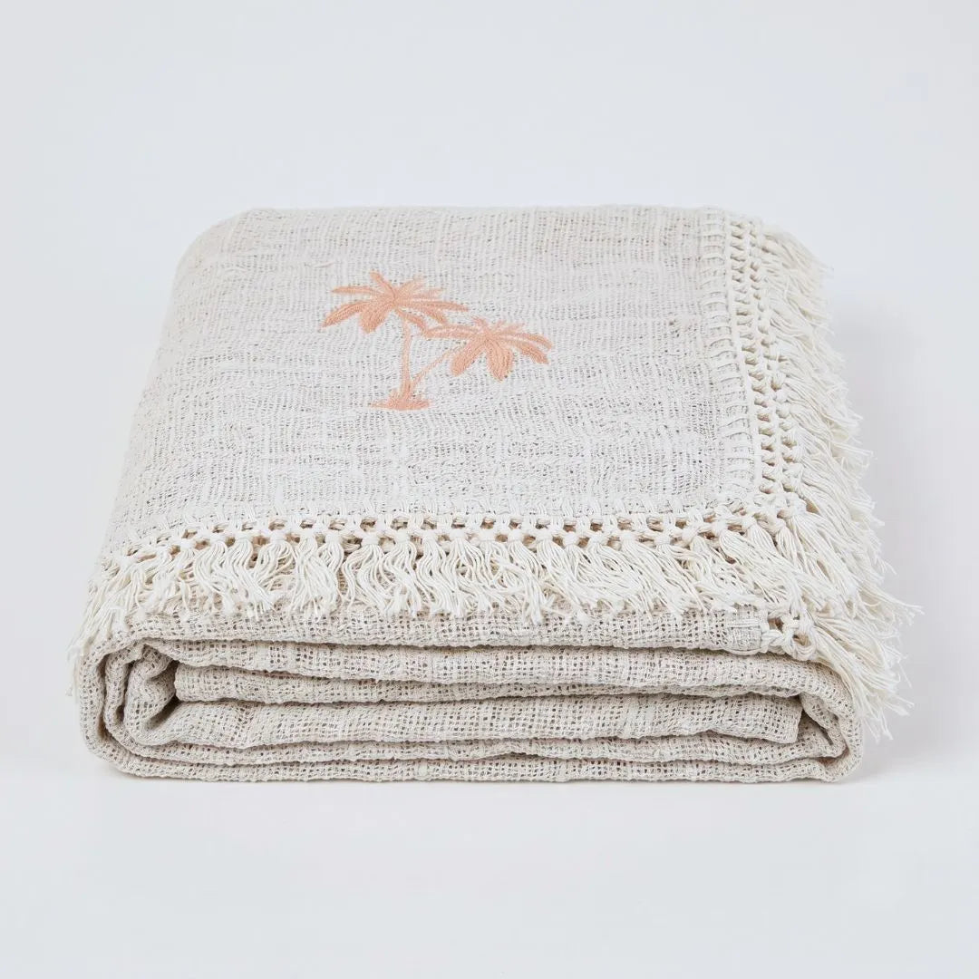 A folded beige linen throw blanket with fringed edges and a small peach palm tree motif, photographed on a clean white background.