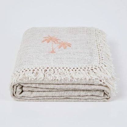 A folded beige linen throw blanket with fringed edges and a small peach palm tree motif, photographed on a clean white background.