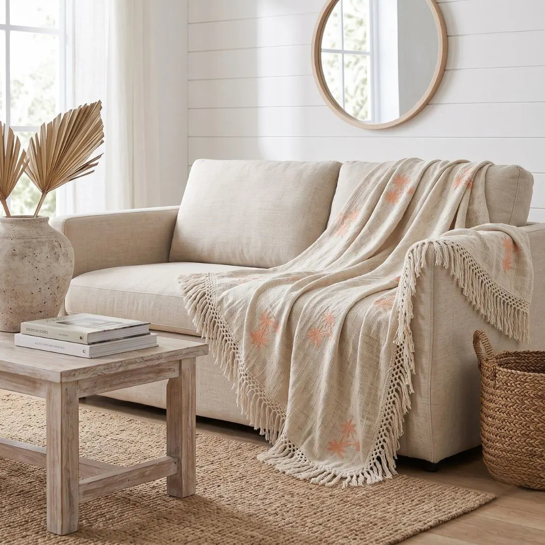 A beige upholstered loveseat in a bright, neutral-toned living room, styled with a cream fringed throw blanket featuring peach floral motifs draped over the seat. A light wood coffee table, woven basket, and round mirror are also visible.