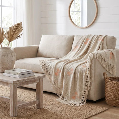 A beige upholstered loveseat in a bright, neutral-toned living room, styled with a cream fringed throw blanket featuring peach floral motifs draped over the seat. A light wood coffee table, woven basket, and round mirror are also visible.