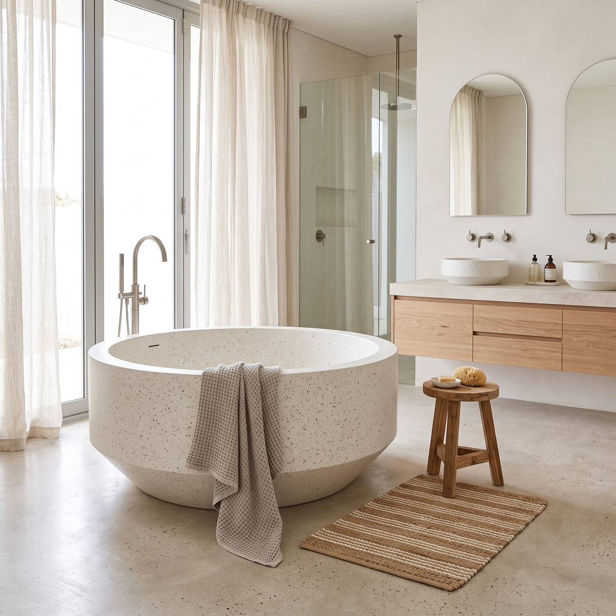 A round freestanding bathtub with a speckled stone-like finish in a bright, modern bathroom. A textured beige towel is draped over the tub, with a woven bath mat and small wooden stool nearby.