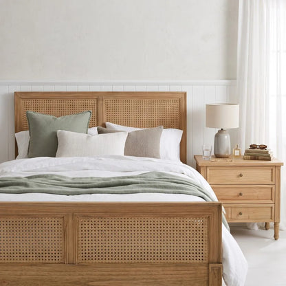 A light natural wood bed frame with woven cane panels, styled with white and sage green bedding, next to a matching wooden nightstand with a lamp.