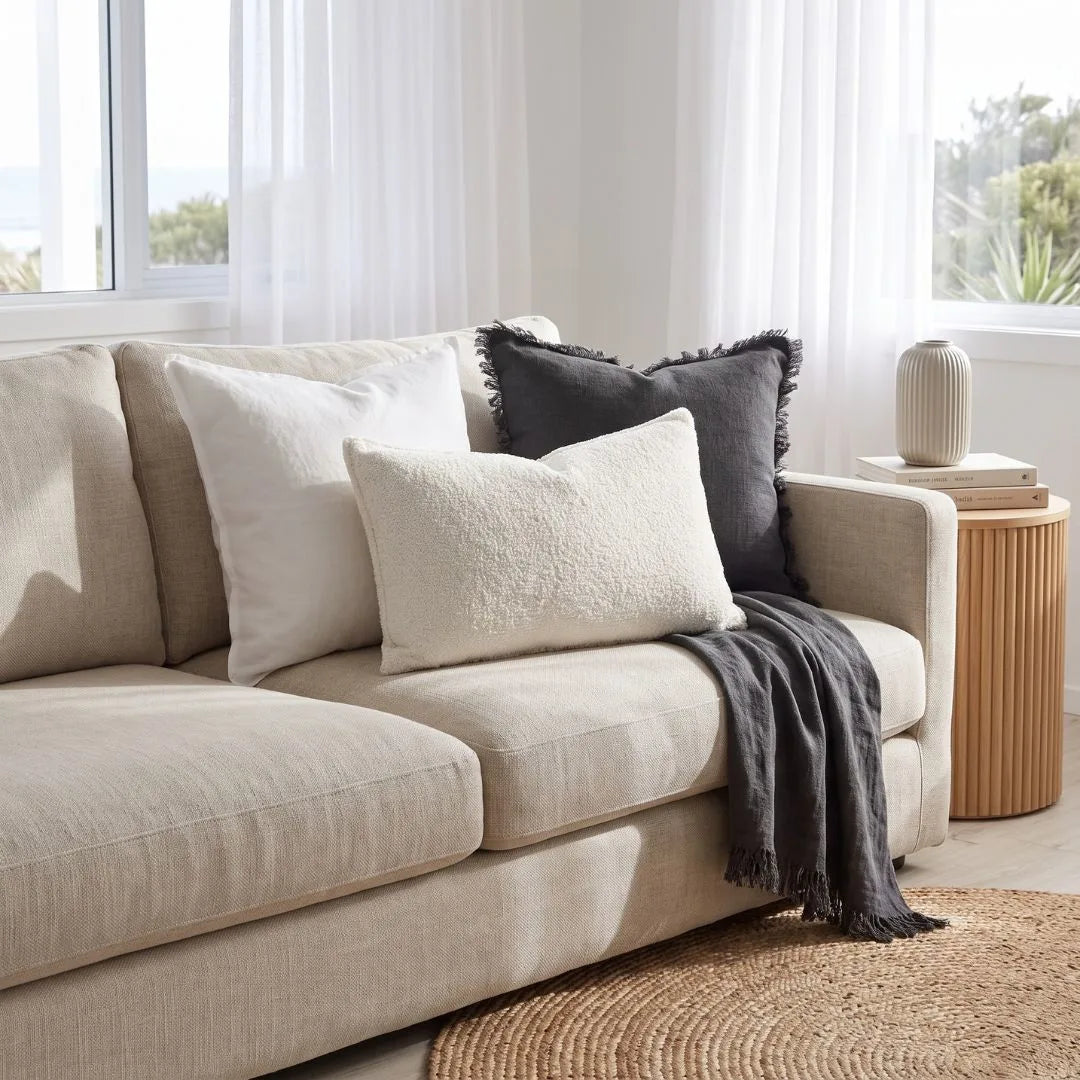 A beige upholstered sofa in a bright living room, styled with three throw pillows in cream and dark charcoal gray, plus a draped dark gray throw blanket. A small round wooden side table with a ribbed vase sits beside the sofa, and a woven round rug is visible in the foreground.