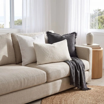 A beige upholstered sofa in a bright living room, styled with three throw pillows in cream and dark charcoal gray, plus a draped dark gray throw blanket. A small round wooden side table with a ribbed vase sits beside the sofa, and a woven round rug is visible in the foreground.