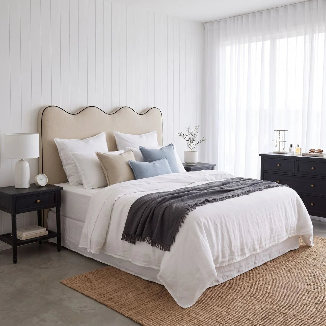 A styled bedroom scene featuring a bed dressed in white linens with beige and light blue pillows, a dark gray throw blanket, a beige scalloped headboard, black nightstands, and a woven rug in a bright, modern room.