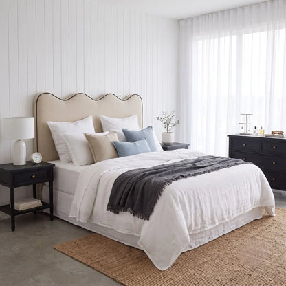 A styled bedroom scene featuring a bed dressed in white linens with beige and light blue pillows, a dark gray throw blanket, a beige scalloped headboard, black nightstands, and a woven rug in a bright, modern room.