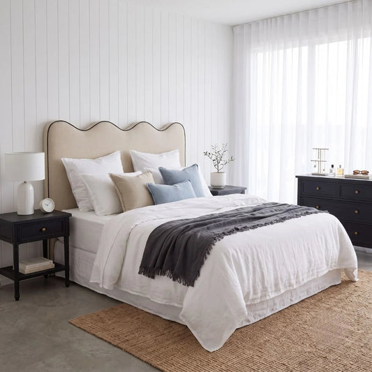 A styled bedroom scene featuring a bed dressed in white linens with beige and light blue pillows, a dark gray throw blanket, a beige scalloped headboard, black nightstands, and a woven rug in a bright, modern room.