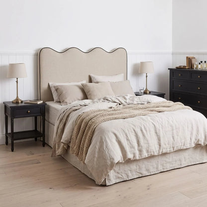 A neatly made bed in a bright bedroom setting, featuring a beige upholstered headboard, neutral beige bedding, matching pillows, a textured throw blanket, a black nightstand on each side with table lamps, and a dark wood dresser against a white paneled wall.