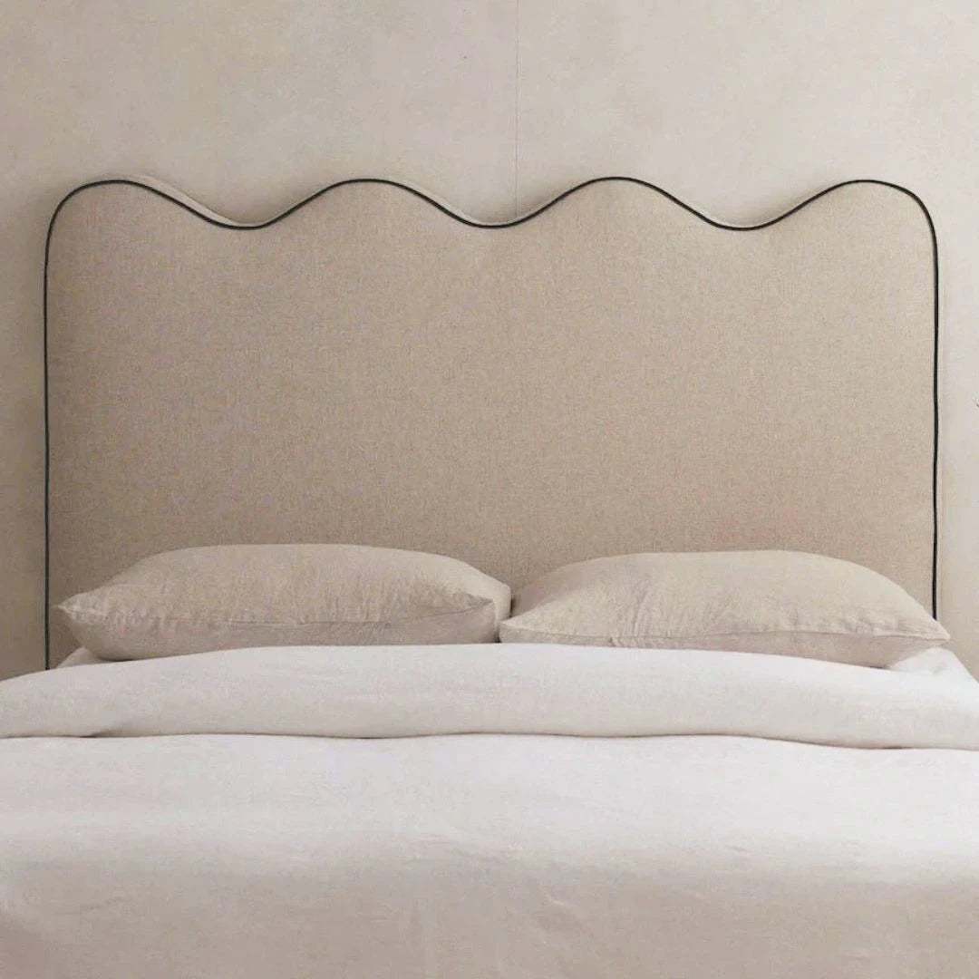 A minimalist beige upholstered headboard with a wavy black metal outline, shown on a bed with white bedding and two pillows against a light beige wall.