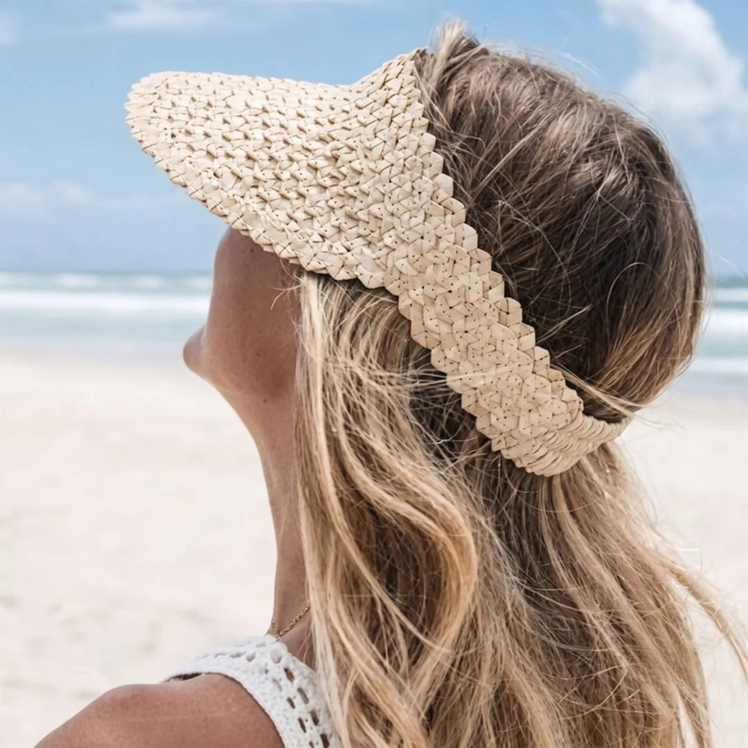 A woman on a beach wearing a woven straw sun visor hat in a light beige color, seen from behind as she looks up toward the sky.