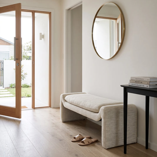 A modern upholstered entryway bench in a light beige fabric, styled in a minimalist entryway with a round gold-framed mirror, light wood flooring, and a black console table.