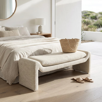 A light beige upholstered entryway bench with a thick seat cushion sits at the foot of a bed in a bright, modern bedroom. A woven straw tote bag rests on the bench, and a pair of beige slide sandals lies on the light wood floor nearby.
