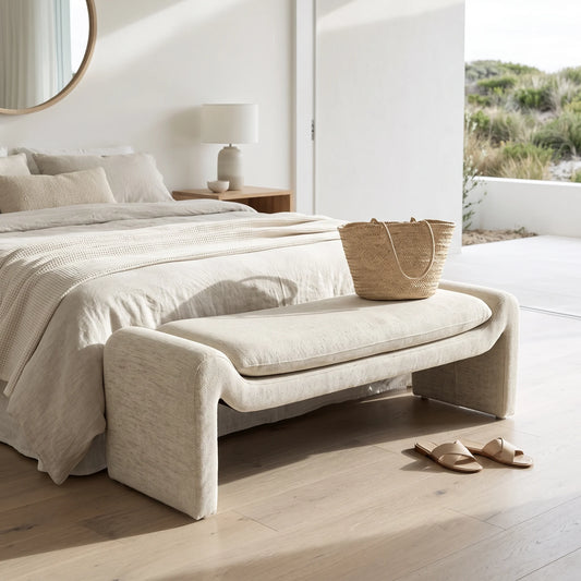 A light beige upholstered entryway bench with a thick seat cushion sits at the foot of a bed in a bright, modern bedroom. A woven straw tote bag rests on the bench, and a pair of beige slide sandals lies on the light wood floor nearby.