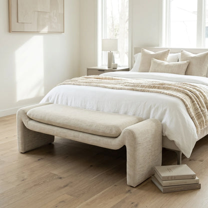 A modern upholstered bed in a bright, minimalist bedroom. The bed is dressed in white linens and a textured beige throw blanket, with a matching beige upholstered bench at the foot of the bed.