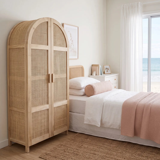 A tall arched wooden wardrobe with woven cane panels stands beside a neatly made bed in a bright, beach-inspired bedroom. The room has white walls, sheer curtains, and a window looking out to the ocean.
