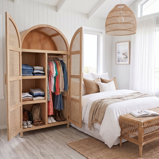 A large arched wooden wardrobe with open doors stands beside a neatly made bed in a bright, white-walled bedroom. The wardrobe has woven cane panels, open shelves with folded clothes, and hanging garments.