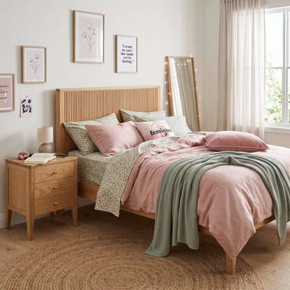 A styled bedroom scene featuring a wooden bed frame with blush pink bedding, patterned green and white duvet covers, a sage green throw blanket, a matching wooden nightstand, and framed wall art.