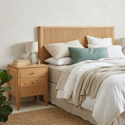 A styled bedroom scene featuring a light natural oak nightstand with three drawers, styled with a table lamp, books, and glasses next to a bed with neutral linens and a green accent pillow.