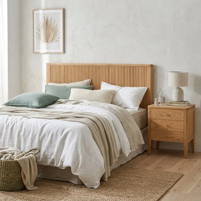 A styled bedroom scene featuring a light natural wood bed frame with a ribbed headboard, dressed in neutral-toned linens, alongside a matching wood nightstand and a textured wall.