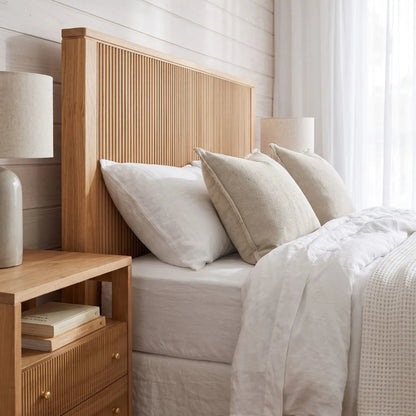 A light natural wood bed frame with a vertically slatted headboard, styled with white and beige bedding, pillows, and a matching wooden nightstand in a bright bedroom.