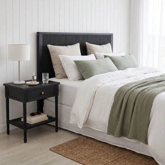 A styled bedroom scene showing a bed with white bedding, sage green pillows and throw, a dark wood headboard and matching nightstand, a white lamp, and a light wood floor.