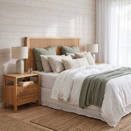 A styled bedroom scene featuring a bed with a light natural wood slatted headboard, dressed in white linens with sage green pillows and a throw blanket. A matching wooden nightstand with a table lamp sits beside the bed against a white shiplap wall.