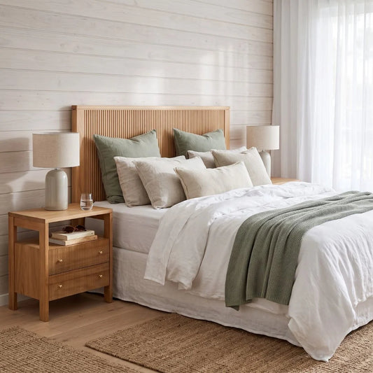 A styled bedroom scene featuring a bed with a light natural wood slatted headboard, dressed in white linens with sage green pillows and a throw blanket. A matching wooden nightstand with a table lamp sits beside the bed against a white shiplap wall.