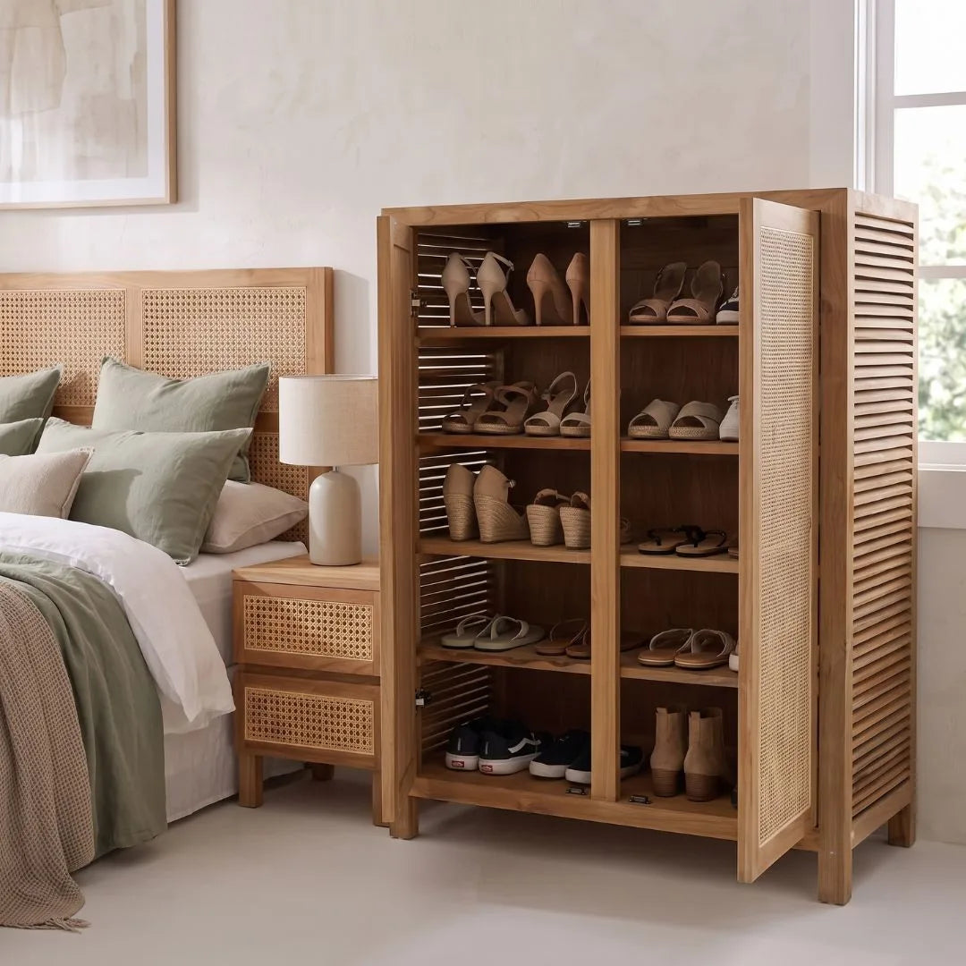 A tall wooden shoe cabinet with open slatted doors and multiple shelves, filled with shoes, placed beside a bed in a softly lit bedroom.