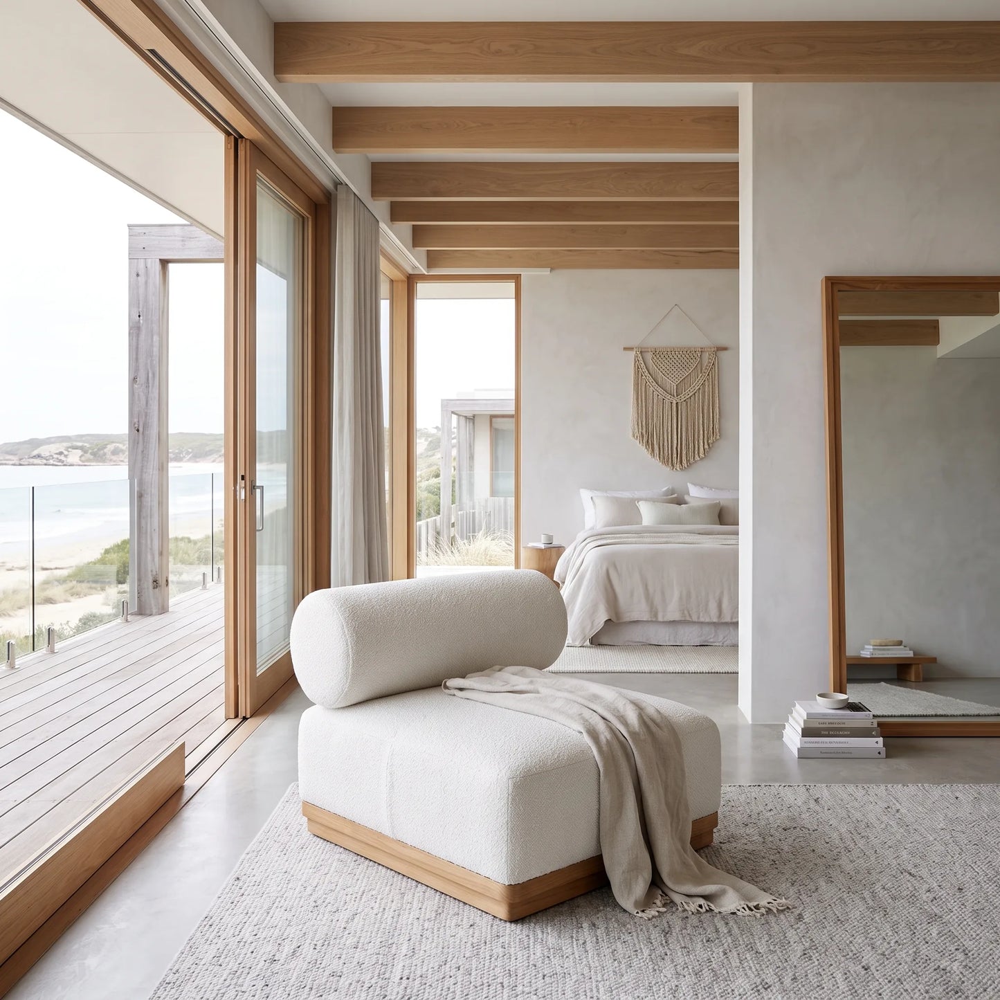 A modern upholstered lounge chair in off-white fabric with a light wood base, styled in a bright, contemporary beachfront bedroom with large windows, wooden beams, and a view of the ocean.